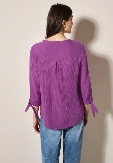 Frau mit lila Bluse und blauer Jeans von hinten vor beigem Hintergrund.